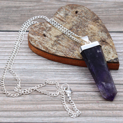Colgante de cristal de color violeta rectangular sobre corazón de madera y cadena plateada en fondo de madera gris