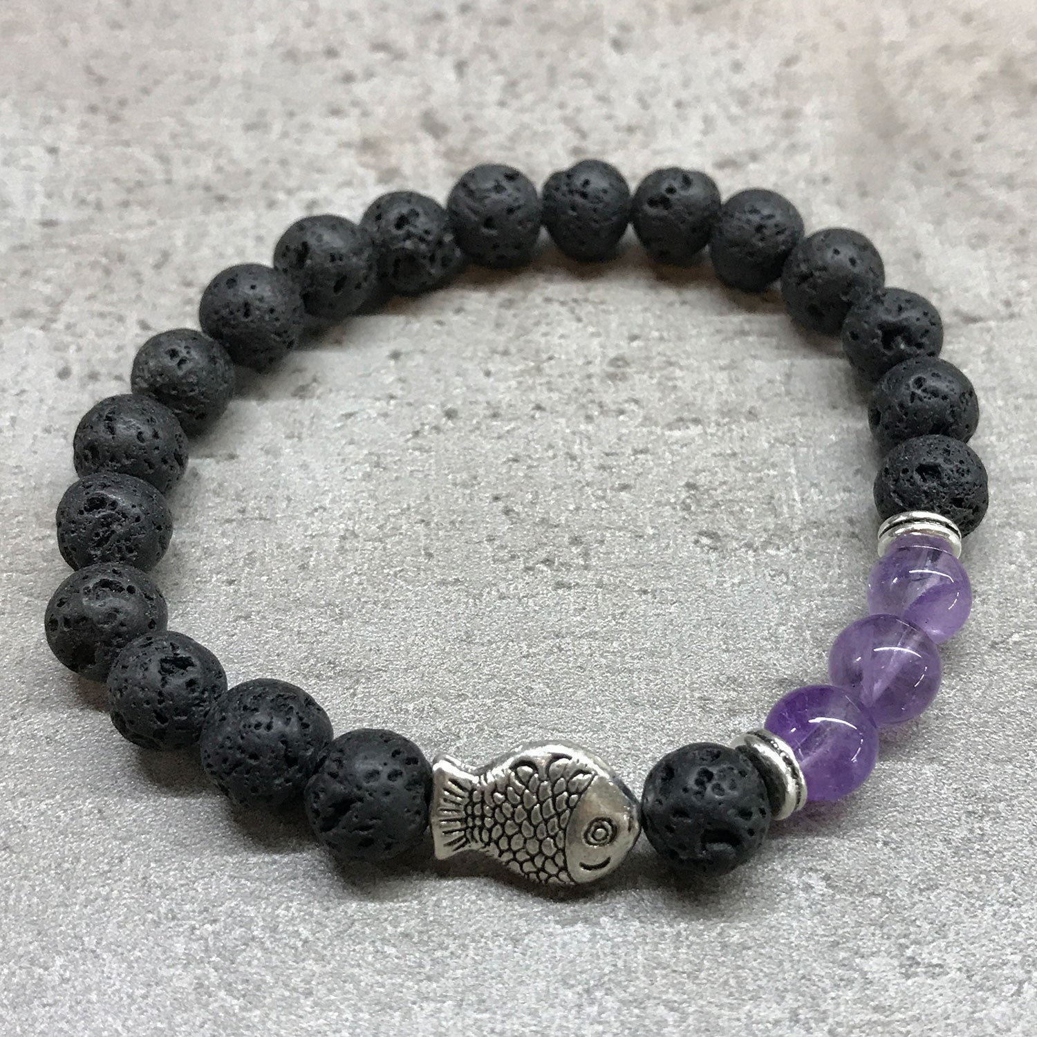 Pulsera con piedras volcánicas negras y 3 piedras de color violeta separadas por una pieza metálica en forma de pez